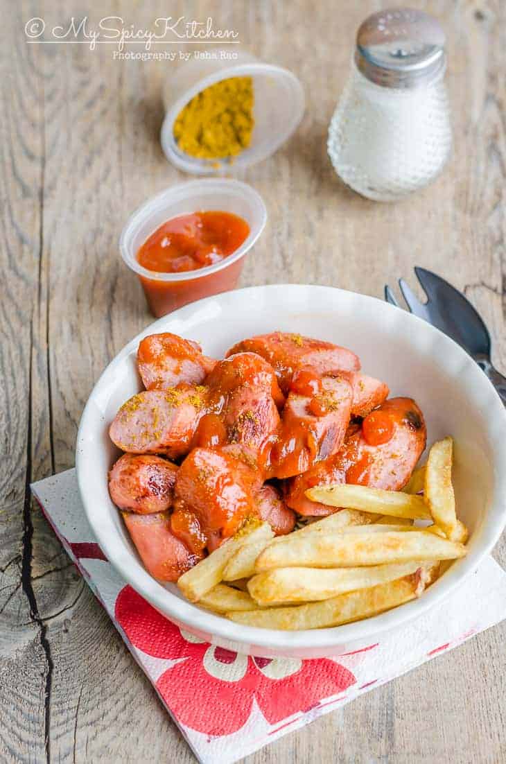 Bowl of currywurst with some fries and, bowl of curry sauce, curry powder and salt on the side.
