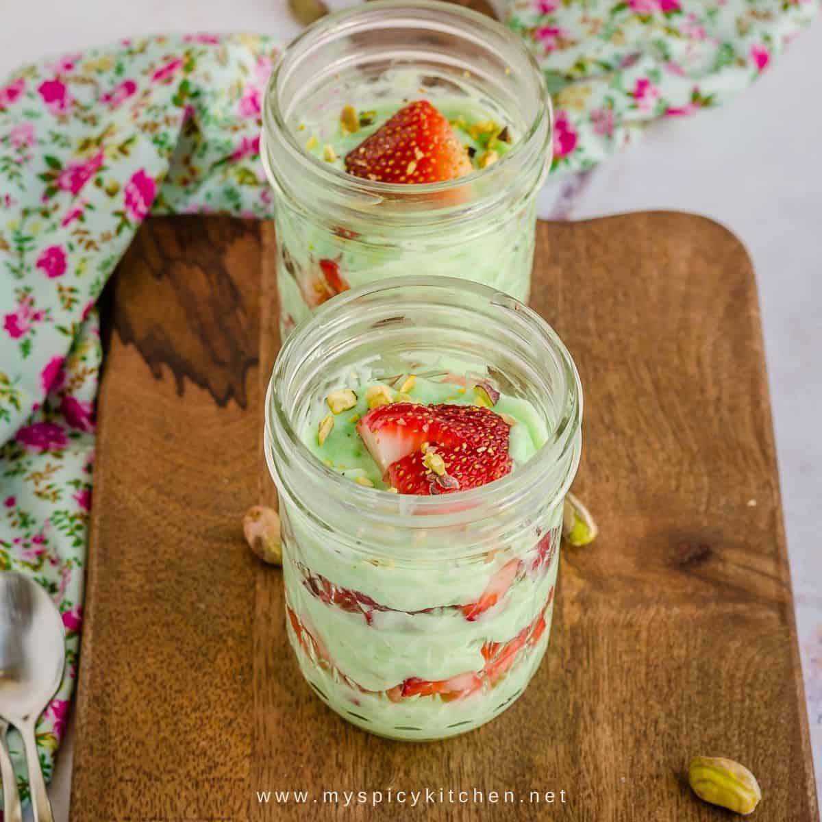 Two jars filled with layers of pistachio pudding and chopped strawberries on a wooden board.