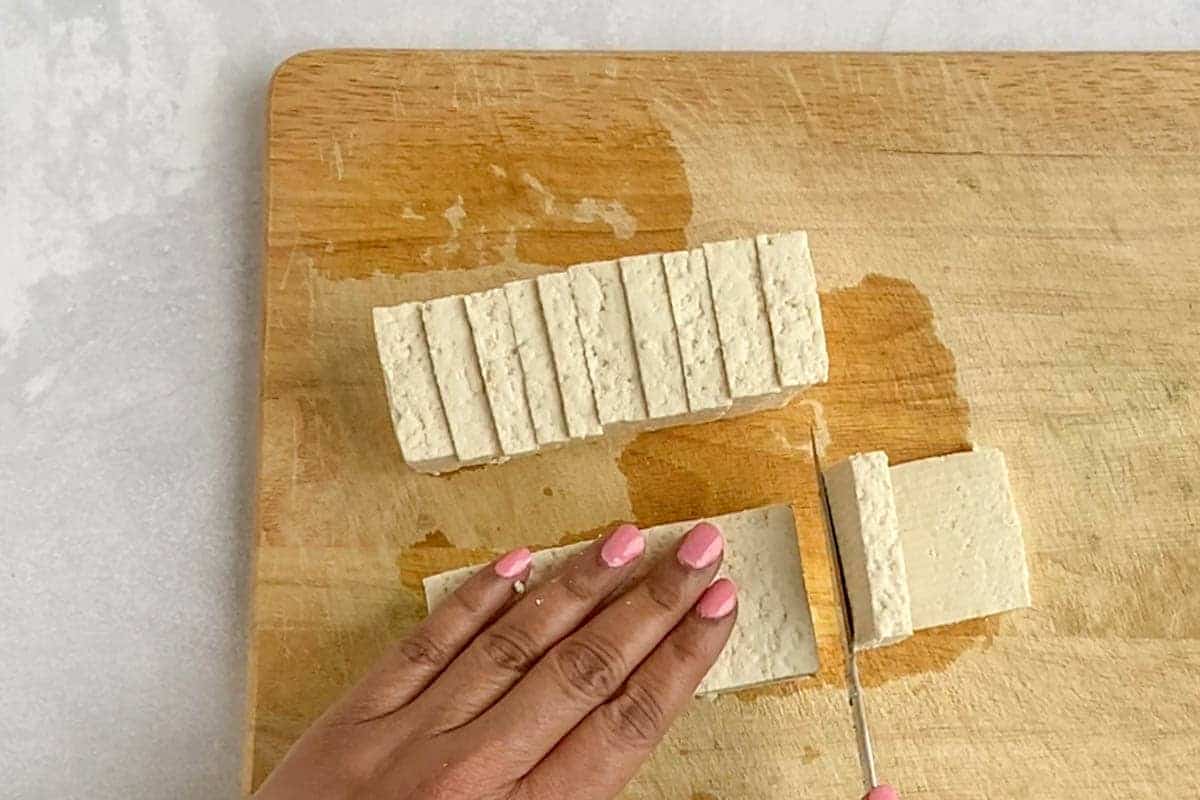 Block of slice tofu and slicing another block of tofu on a wooden cutting board.
