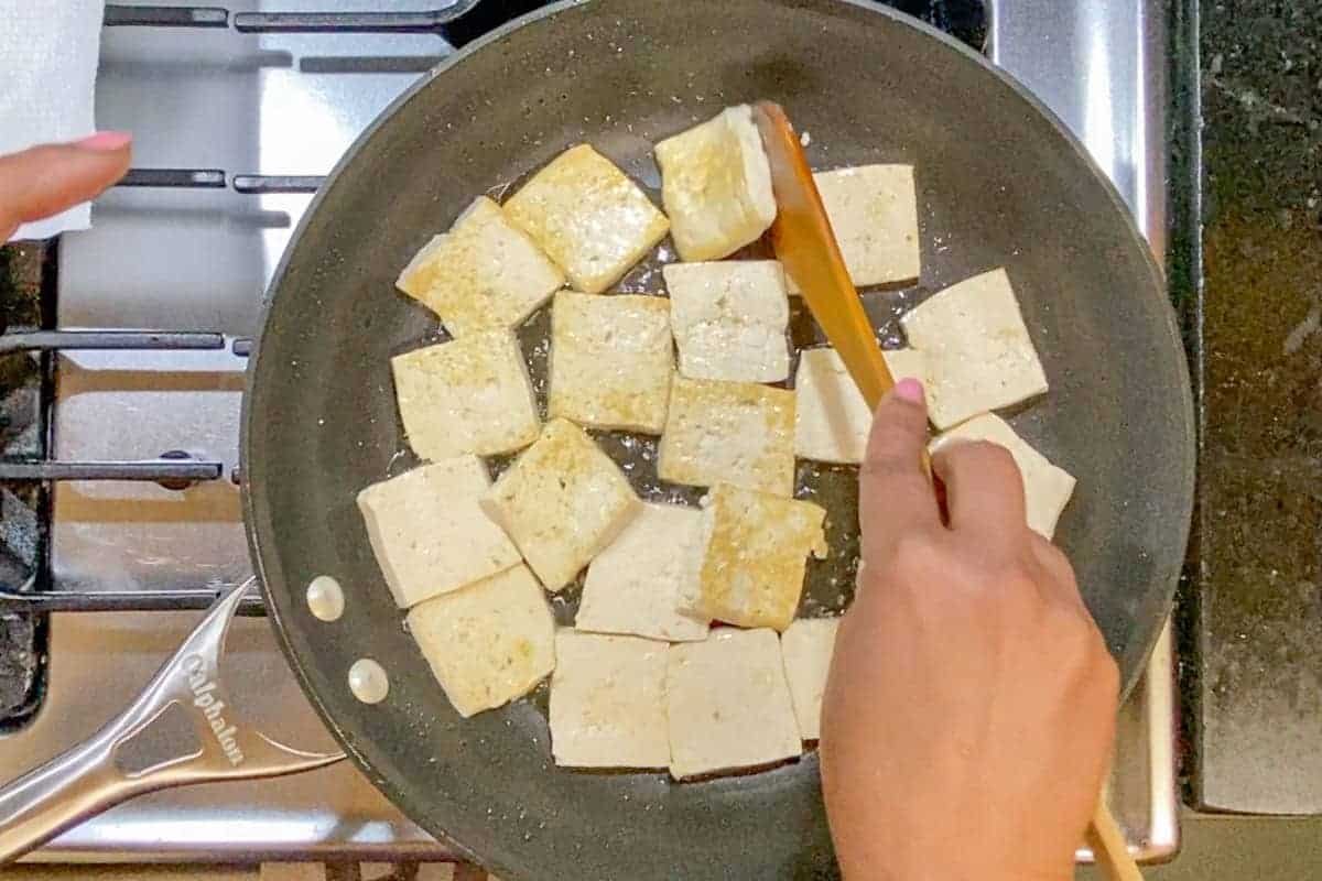 Pan frying tofu in a wide pan and flipping tofu with a wooden spatula.