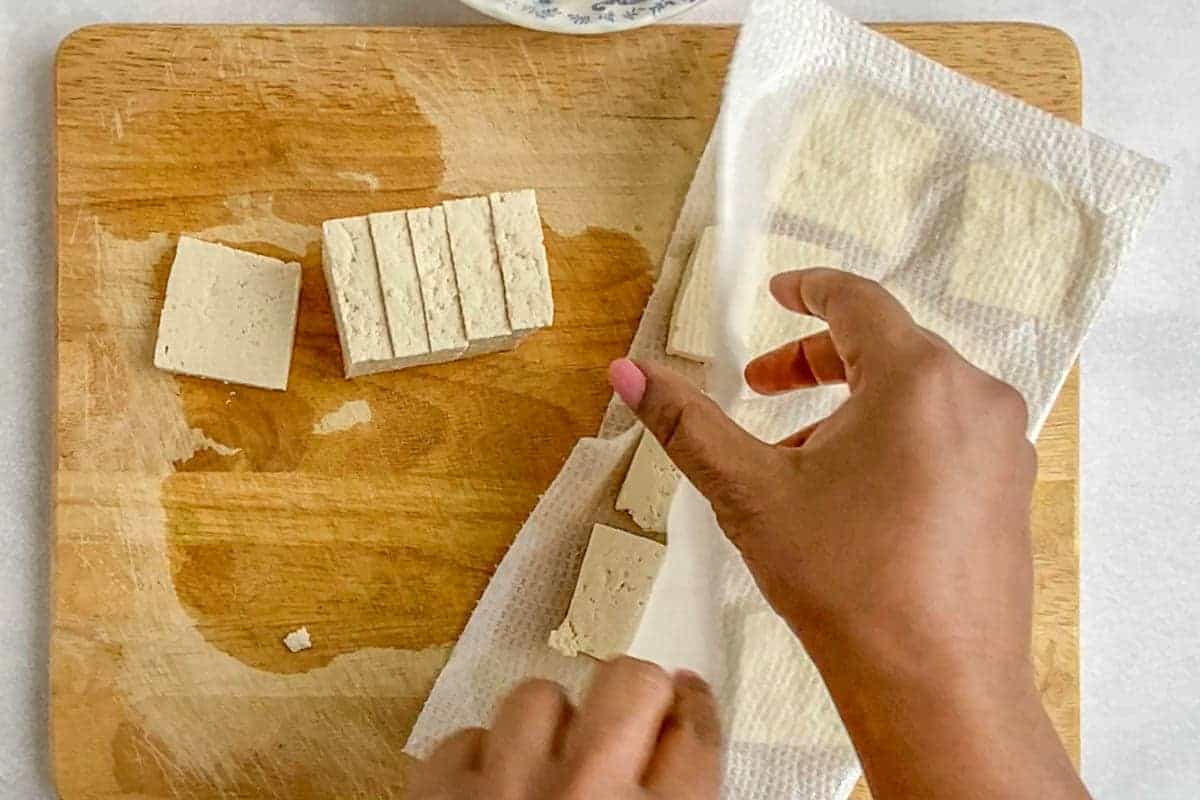 Few slices tofu on a wooden cutting board and pat drying few more slices between two paper towels.